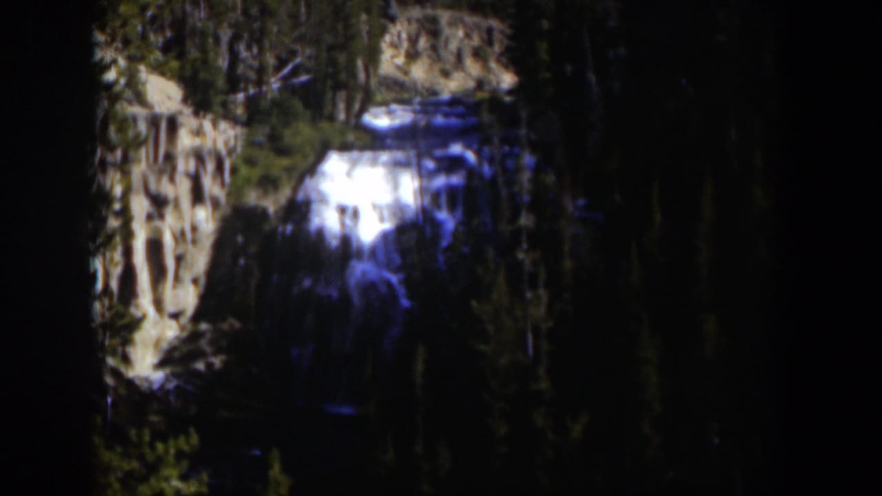 1951 Wyoming 8mm frame showing a bright cascade spilling over layered brown rocks in a dense evergreen setting.