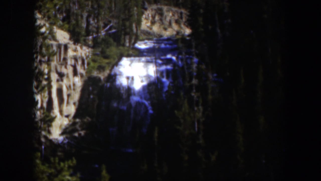 1951 Wyoming 8mm frame showing a bright waterfall framed by evergreens and canyon rock in strong sun and shadow.