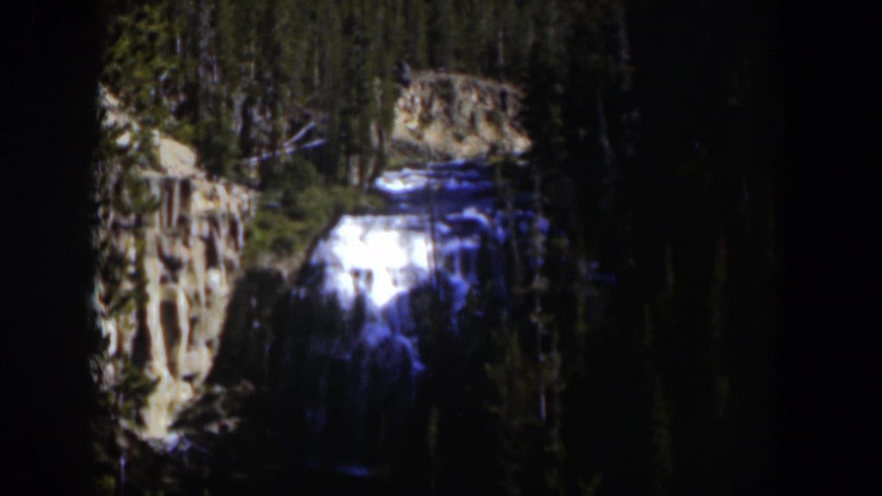 1951 Wyoming 8mm frame of a sunlit waterfall cutting through a shadowed evergreen canyon.