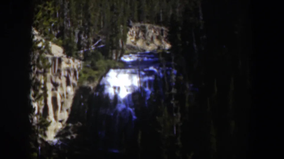 1951 Wyoming 8mm frame of a sunlit waterfall cutting through a shadowed evergreen canyon.