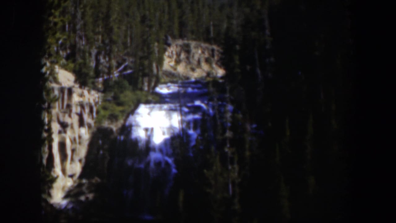 1951 Wyoming 8mm frame showing a distant waterfall amid evergreens, with pale rock and deep shadows around the falls.