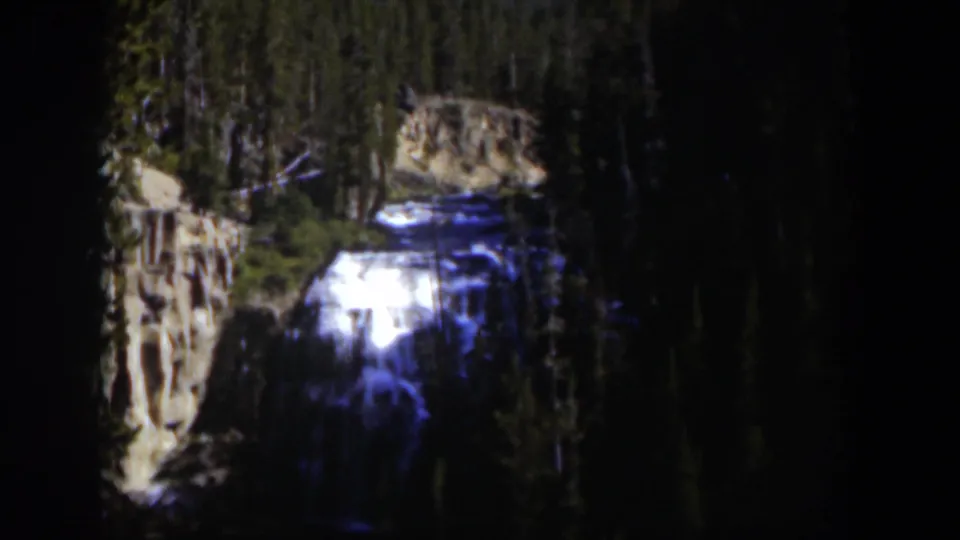1951 Wyoming 8mm frame showing a distant waterfall amid evergreens, with pale rock and deep shadows around the falls.