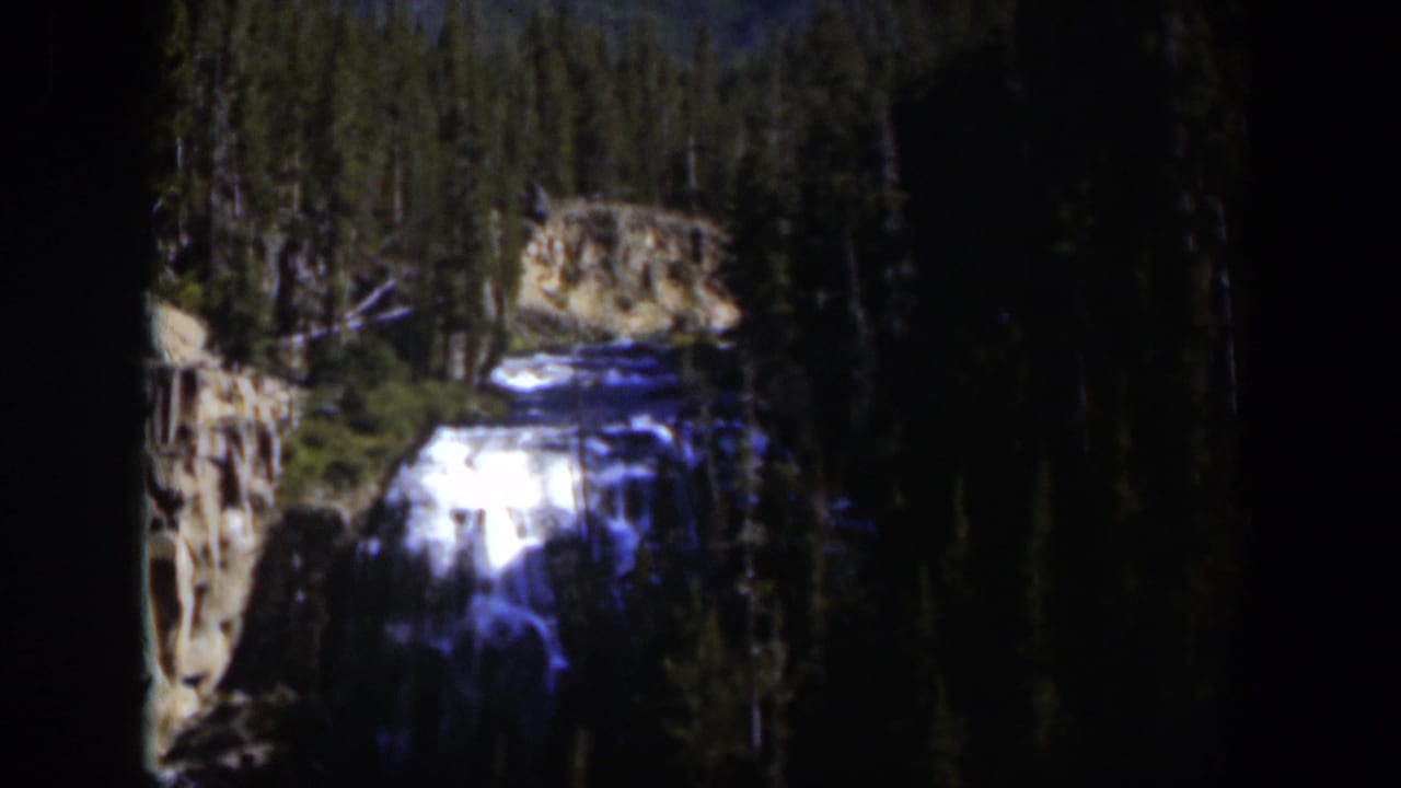 1951 Wyoming 8mm frame showing a bright waterfall descending through a dark evergreen mountain landscape.