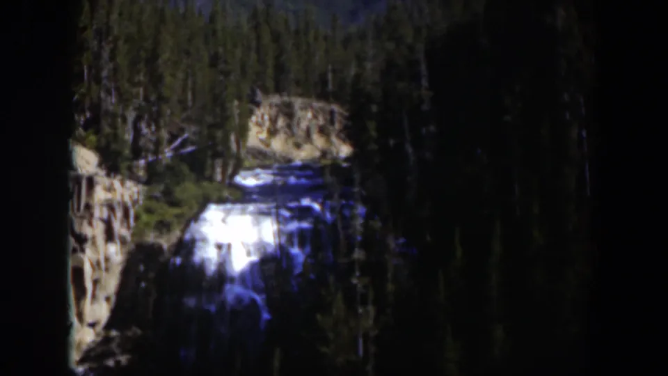 1951 Wyoming 8mm frame showing a bright waterfall descending through a dark evergreen mountain landscape.