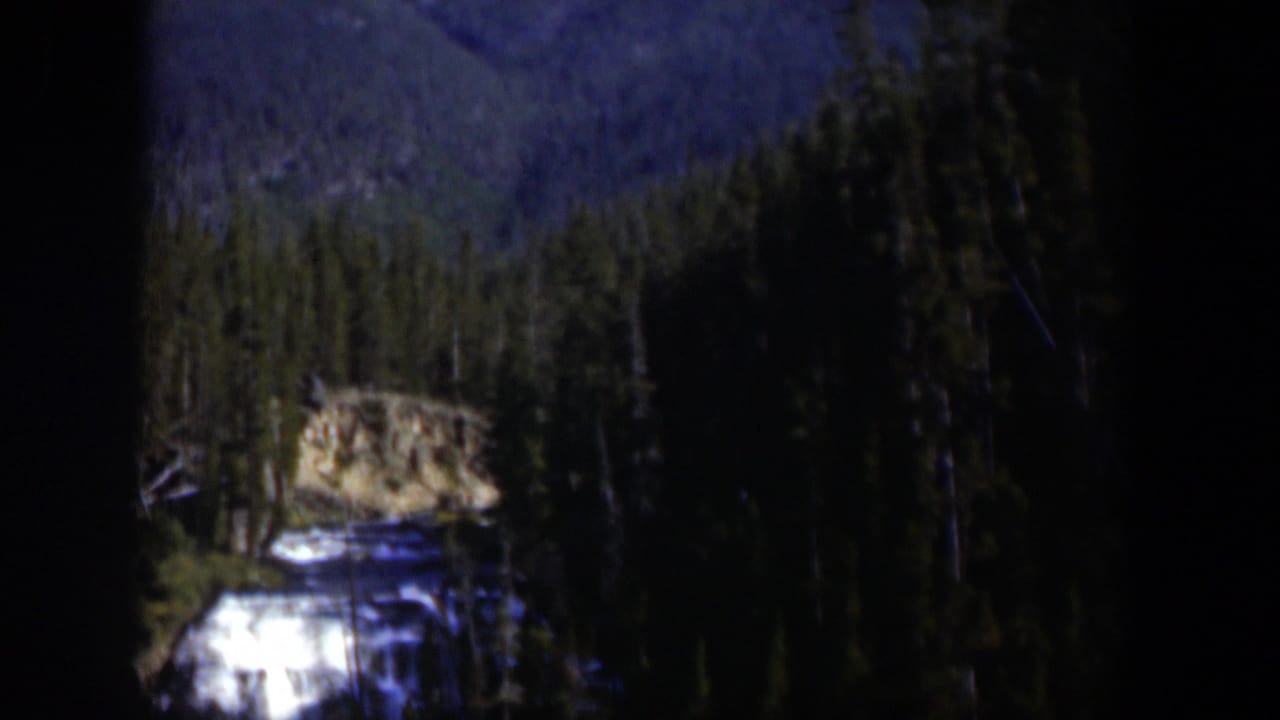 1951 Wyoming 8mm frame showing a small mountain waterfall beneath evergreens and a blue-toned distant slope.
