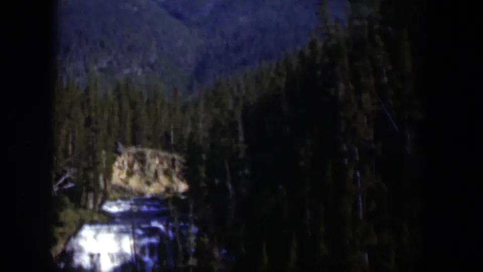 1951 Wyoming 8mm frame showing a small mountain waterfall beneath evergreens and a blue-toned distant slope.