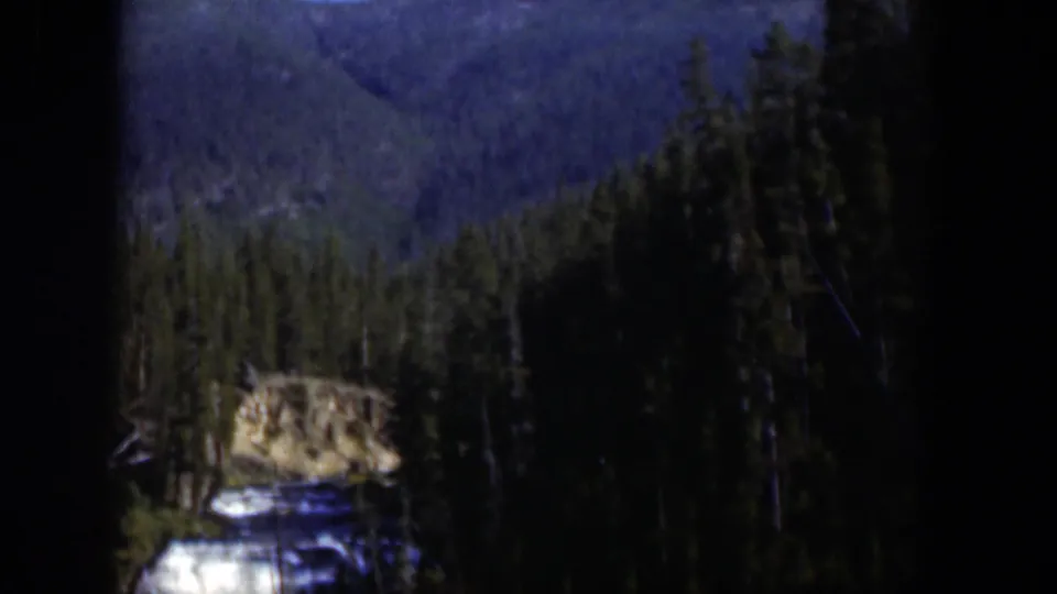 1951 Wyoming 8mm frame showing evergreen forest, blue ridges, and a rocky roadside feature through a dark window frame.