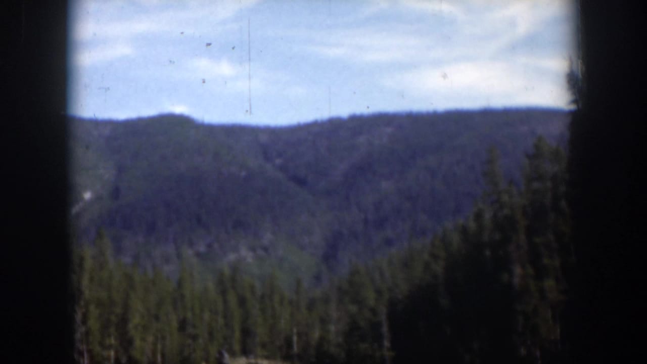 1951 Wyoming 8mm frame with a soft view of forested mountains, a pale blue sky, and dark vertical gate edges.
