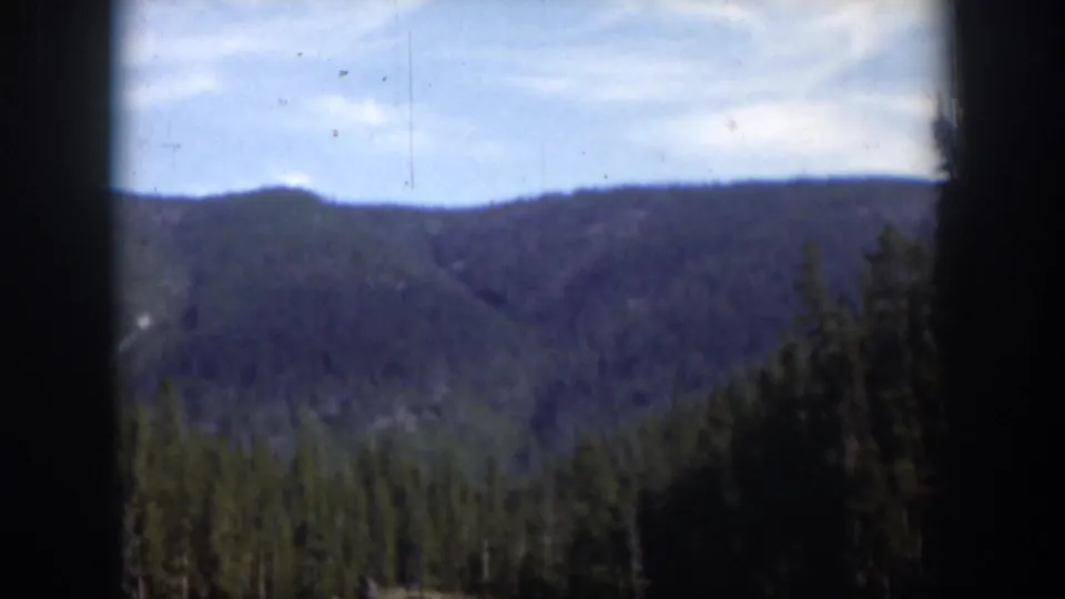1951 Wyoming 8mm frame with a soft view of forested mountains, a pale blue sky, and dark vertical gate edges.