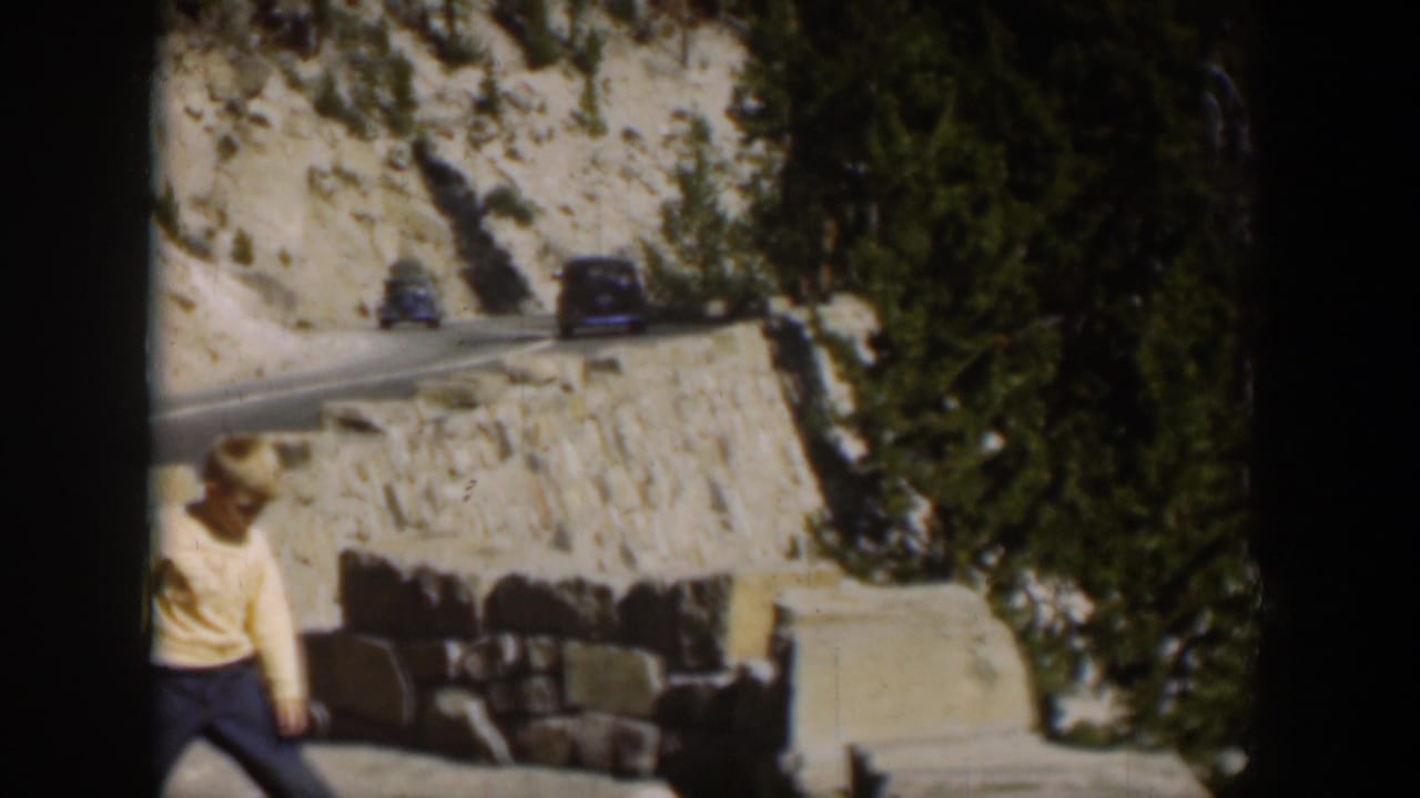 Two cars share the winding mountain road as a boy in a light shirt stands near a roadside wall in Wyoming, 1951.