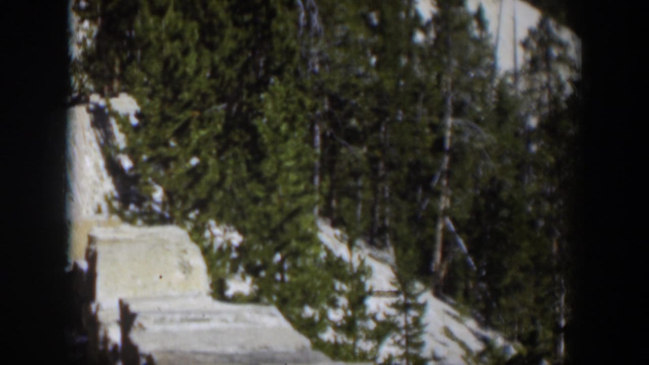 1951 Wyoming 8mm frame with snow, conifers, and a pale wall or road edge crossing a sunlit mountain scene.