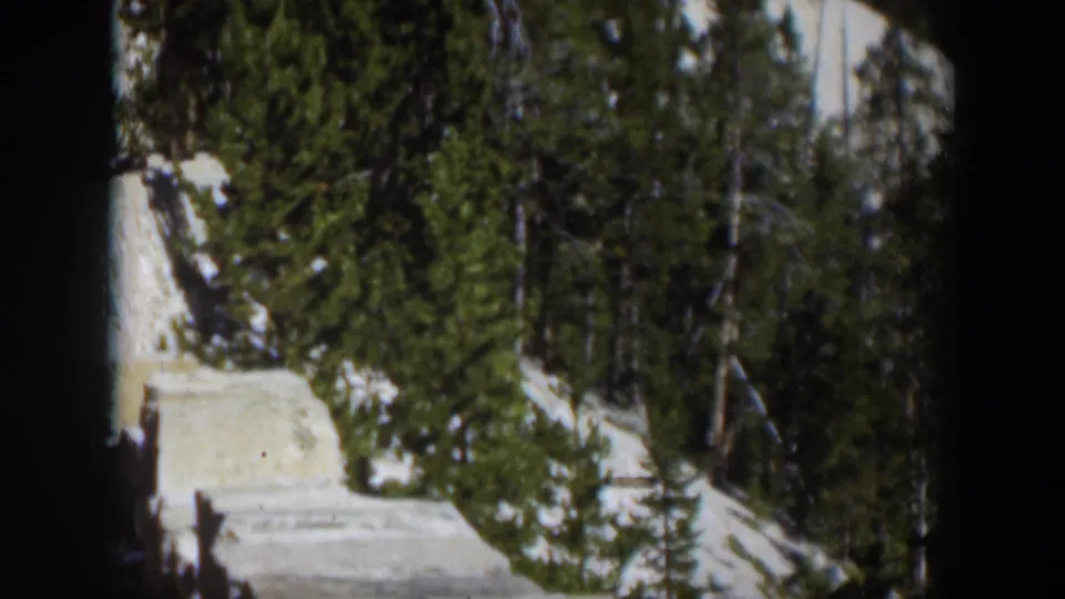 1951 Wyoming 8mm frame with snow, conifers, and a pale wall or road edge crossing a sunlit mountain scene.