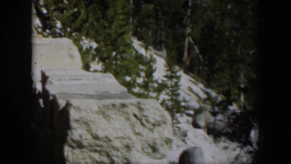1951 Wyoming 8mm still showing a pale rock ledge, dark evergreen trees, and remaining snow on a sunlit slope.