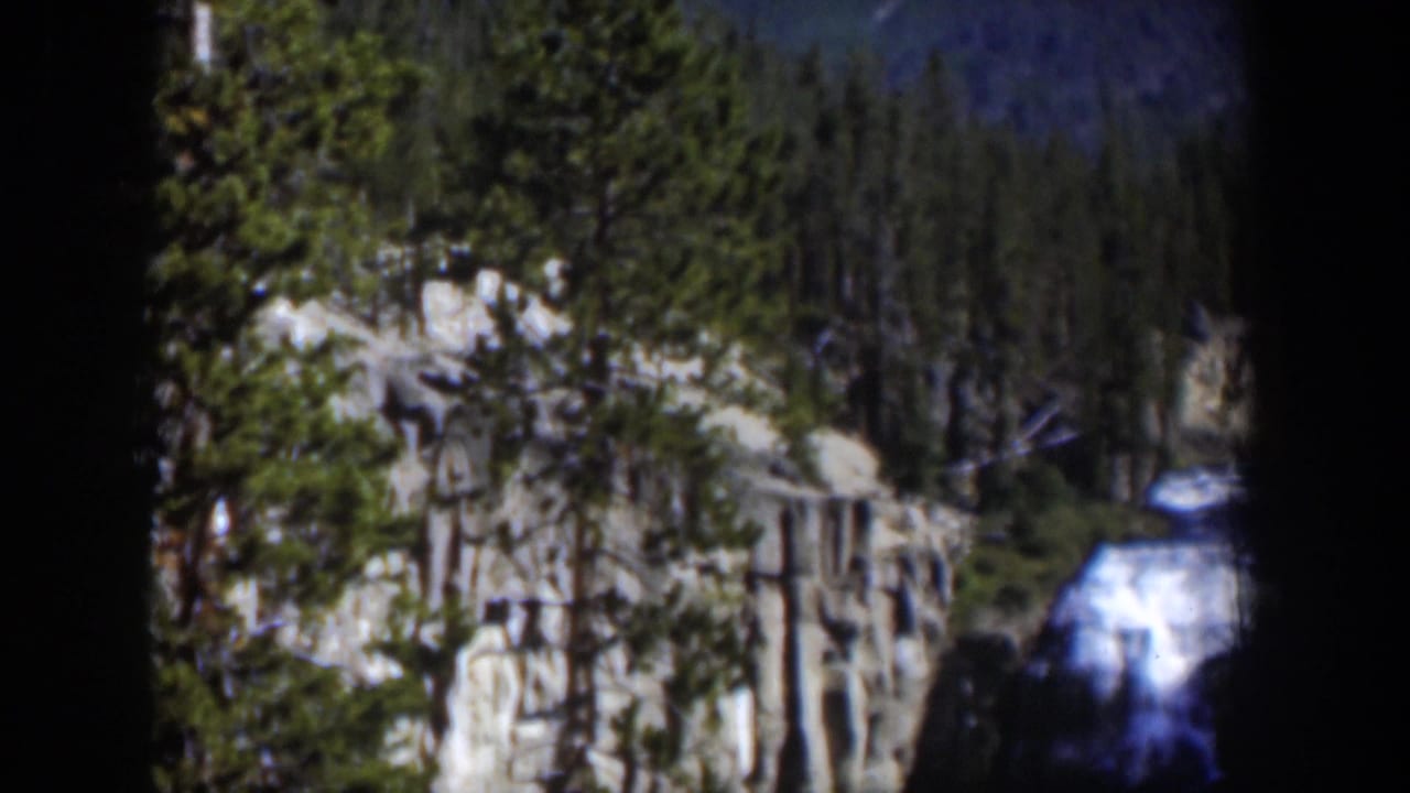 1951 Wyoming home-movie frame showing a waterfall below pale rock cliffs and evergreen trees in dim blue-tinged light.