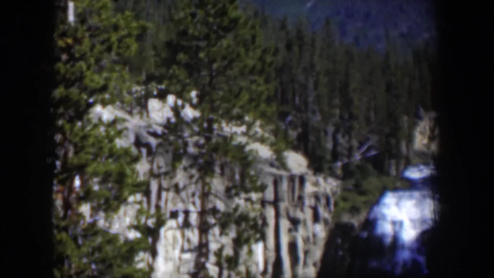 1951 Wyoming home-movie frame showing a waterfall below pale rock cliffs and evergreen trees in dim blue-tinged light.