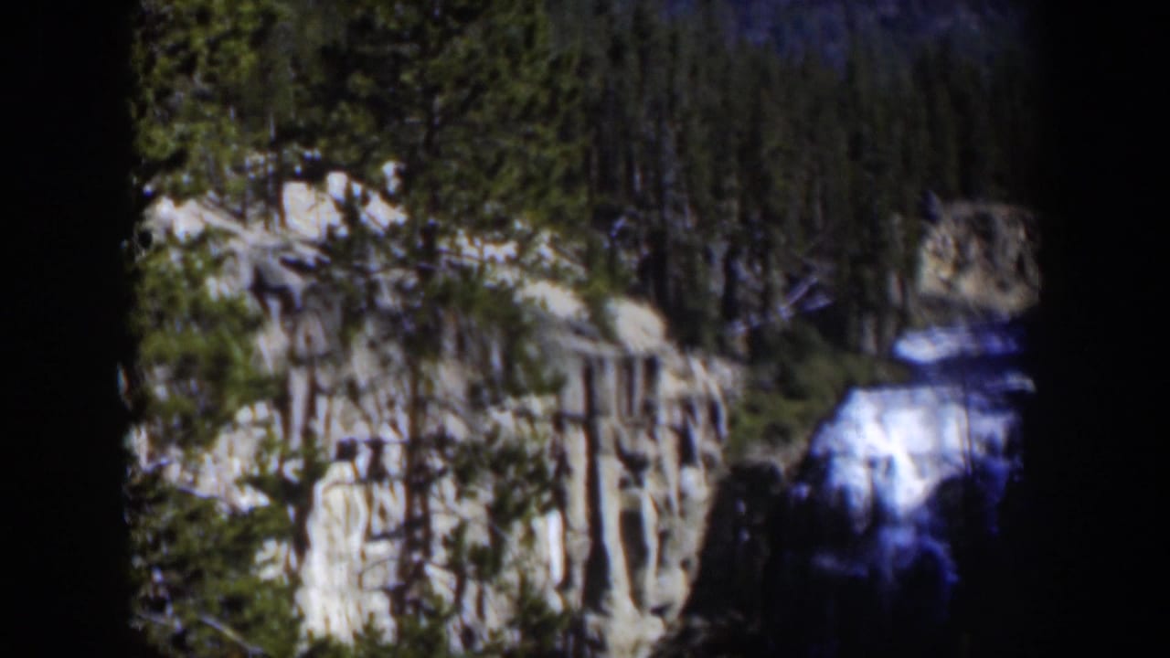 Distant waterfall and rocky cliff with evergreen trees under dark borders.
