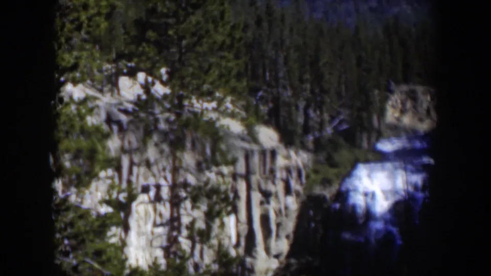 Blue-lit waterfall drops beside a pale cliff and dense trees in this supported 1951 Wyoming 8mm home-movie frame.