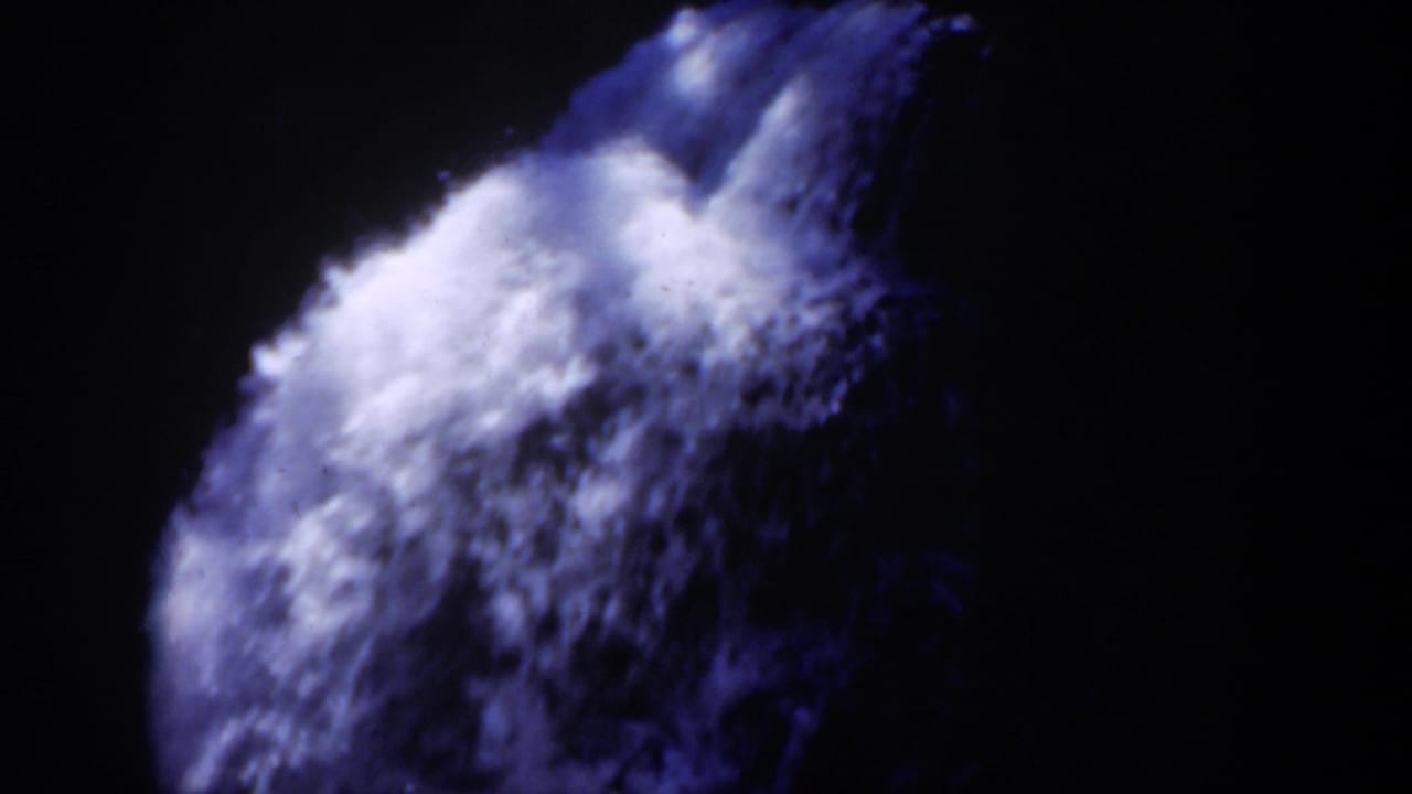 1951 Wyoming 8mm frame showing a bright rush of waterfall foam dropping over shadowed rock, with blue-purple film color and deep surrounding darkness.