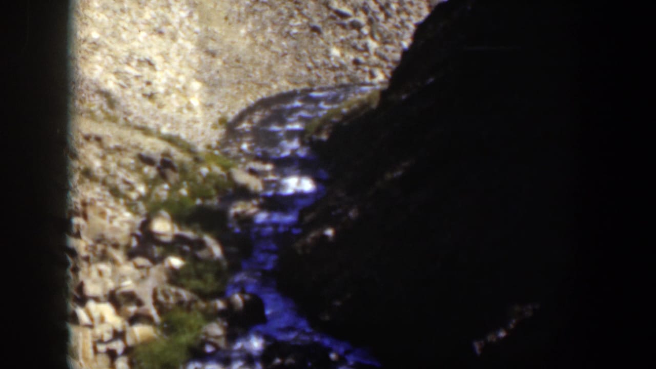 1951 Wyoming home-movie frame showing a narrow stream crossing pale gravel beside dark canyon rock.
