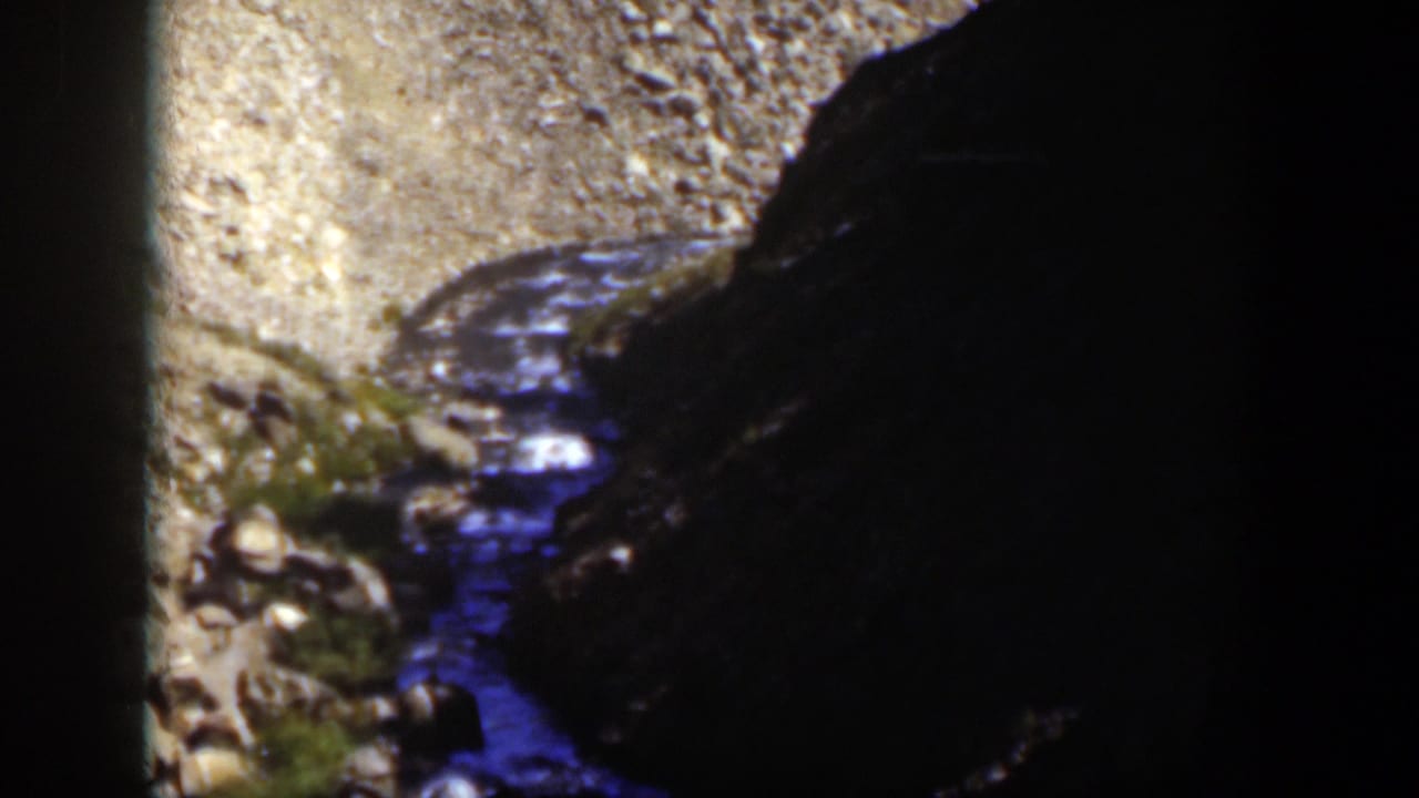 1951 Wyoming 8mm frame of a rocky stream channel bordered by gravel and a large shadowed canyon wall.