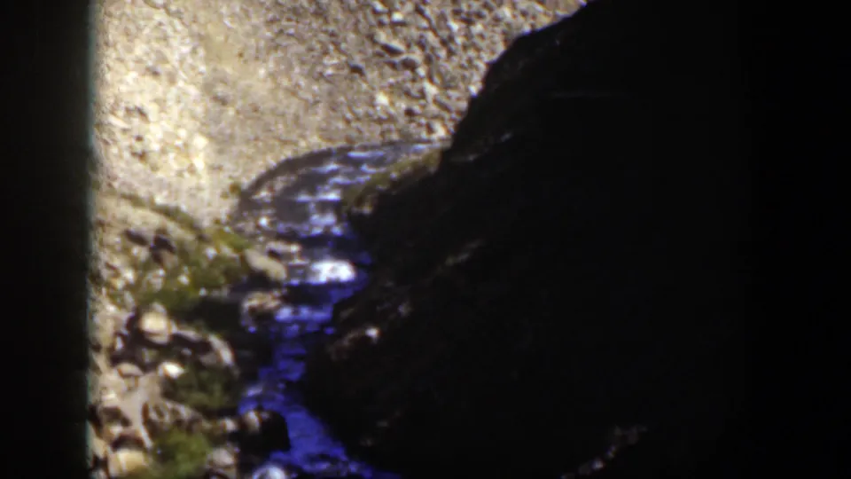 1951 Wyoming 8mm frame of a rocky stream channel bordered by gravel and a large shadowed canyon wall.