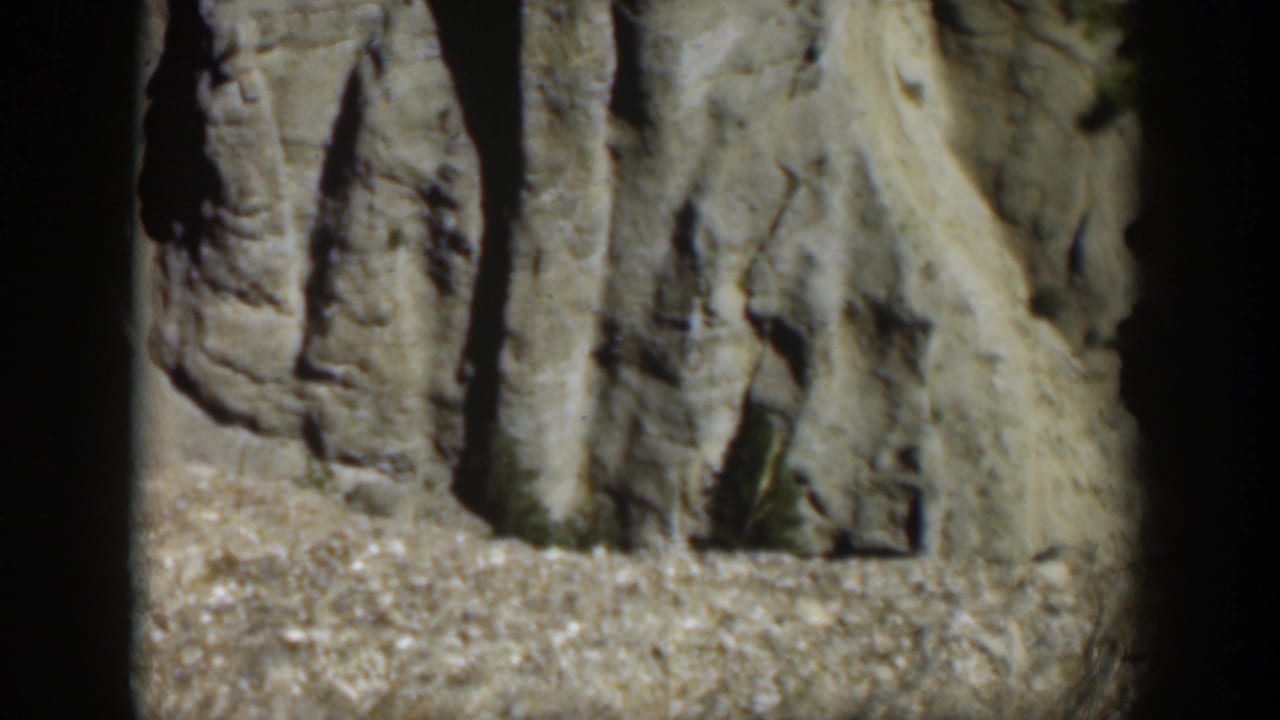 1951 Wyoming frame showing a pale rock cliff face above a gravel slope, framed by dark canyon shadow.