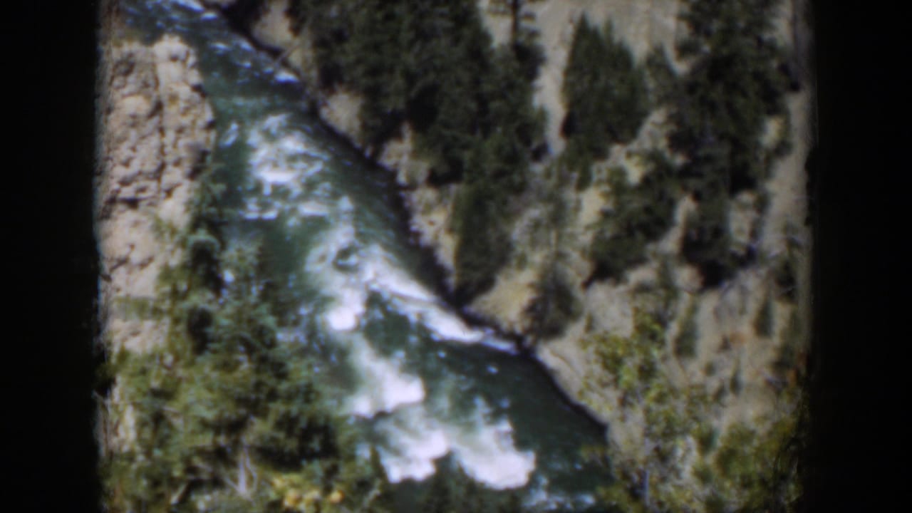 1951 Wyoming 8mm frame of a white-water river below pale canyon rock and evergreen vegetation.