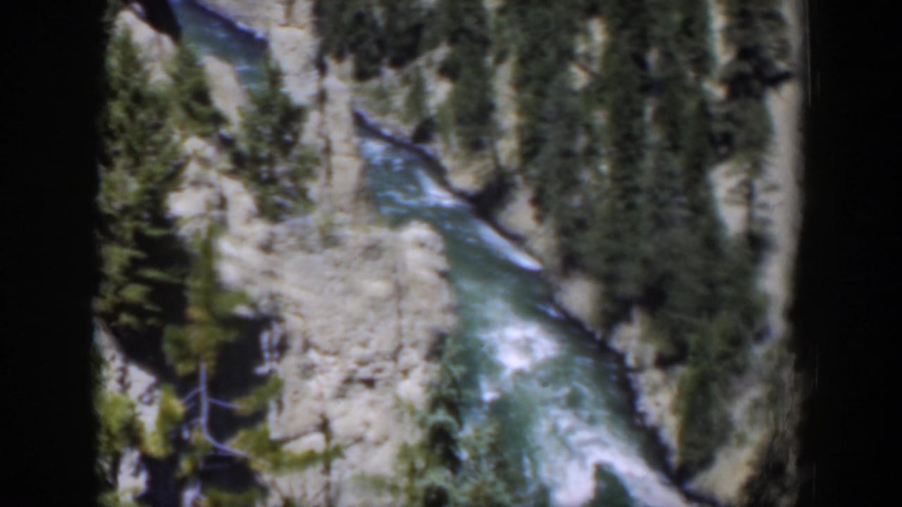 High-angle 1951 Wyoming home-movie view of rushing river water bordered by light rock and sparse pine growth.