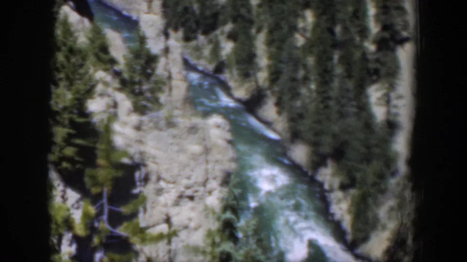 High-angle 1951 Wyoming home-movie view of rushing river water bordered by light rock and sparse pine growth.