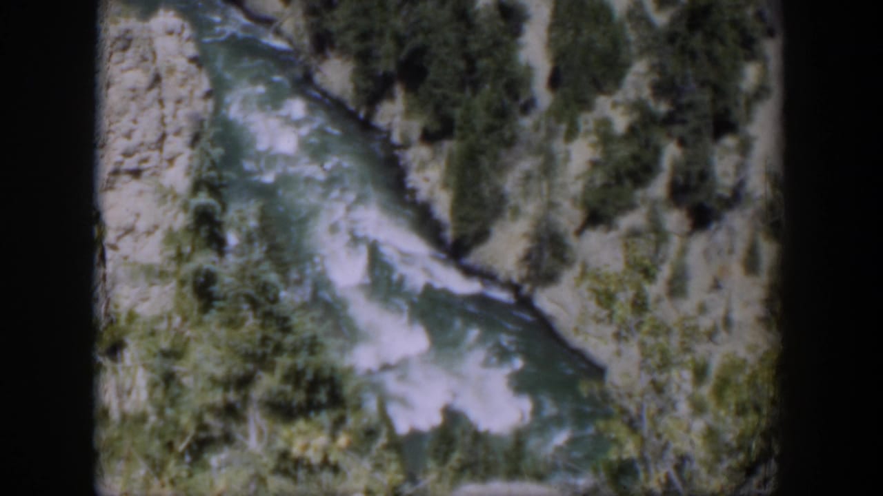 Final 1951 Wyoming frame holds on fast-moving river rapids below rocky canyon banks with scattered pines.