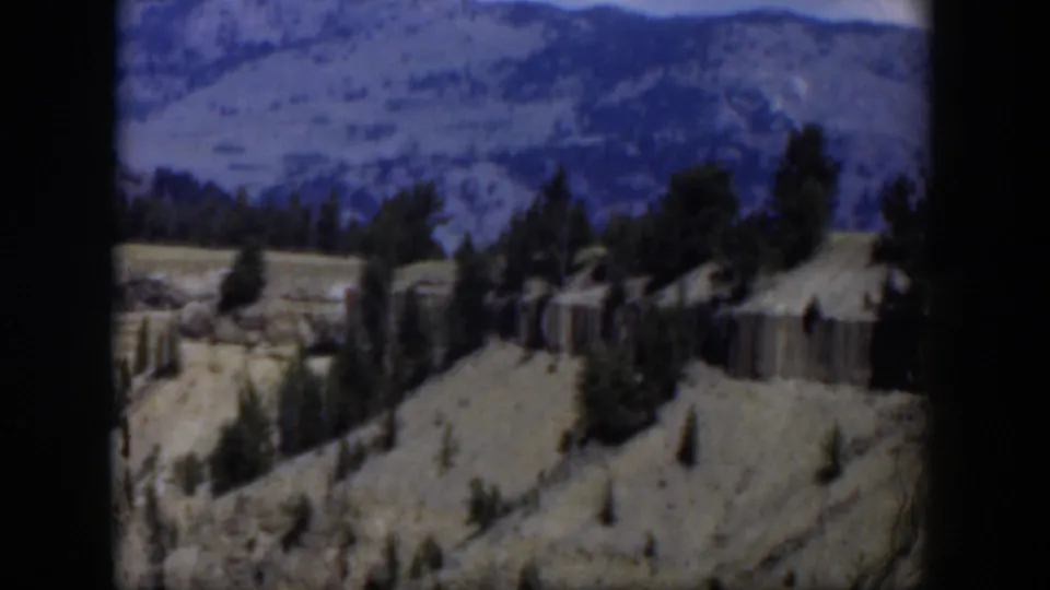 1951 8mm Wyoming home-movie frame showing loose scree, pines, a cliff band, and blue-gray mountains beyond.