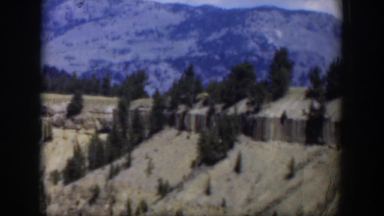 1951 Wyoming landscape frame with a sheer rocky band, scattered pines, loose debris, and mountains in the background.