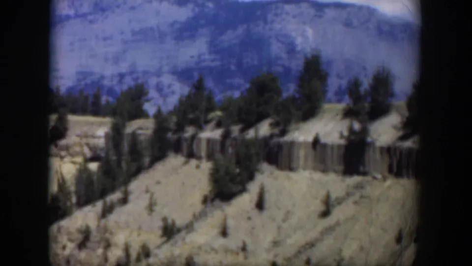 1951 8mm Wyoming frame showing scattered evergreens across a light rocky hillside beneath blue-gray mountains.