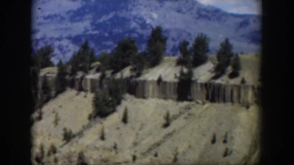 1951 Wyoming 8mm frame showing pale eroded hills, pine trees, a cliff band, and distant blue mountains from an aerial viewpoint.