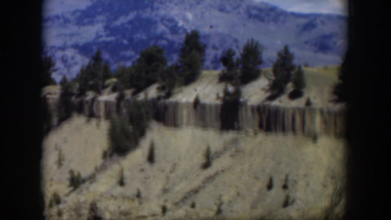 1951 Wyoming aerial frame with a horizontal cliff face, sparse pine trees, rolling tan terrain, and blue mountain ridges beyond.