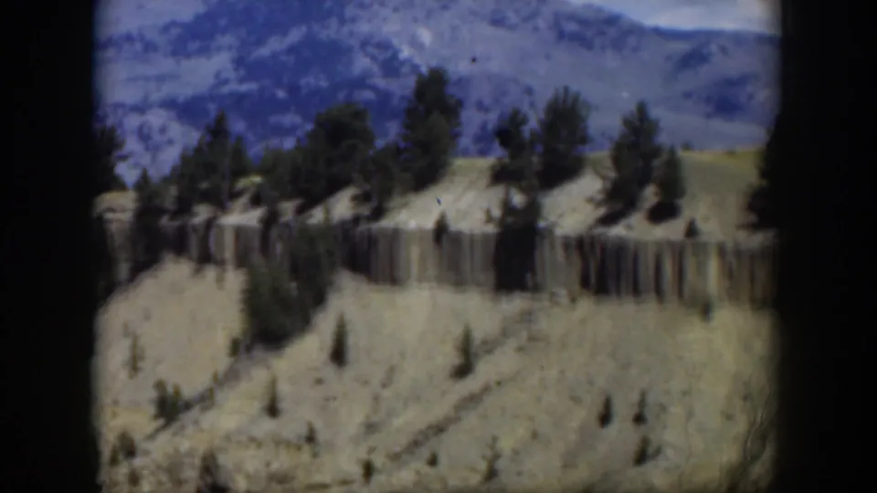 1951 Wyoming aerial frame with a horizontal cliff face, sparse pine trees, rolling tan terrain, and blue mountain ridges beyond.