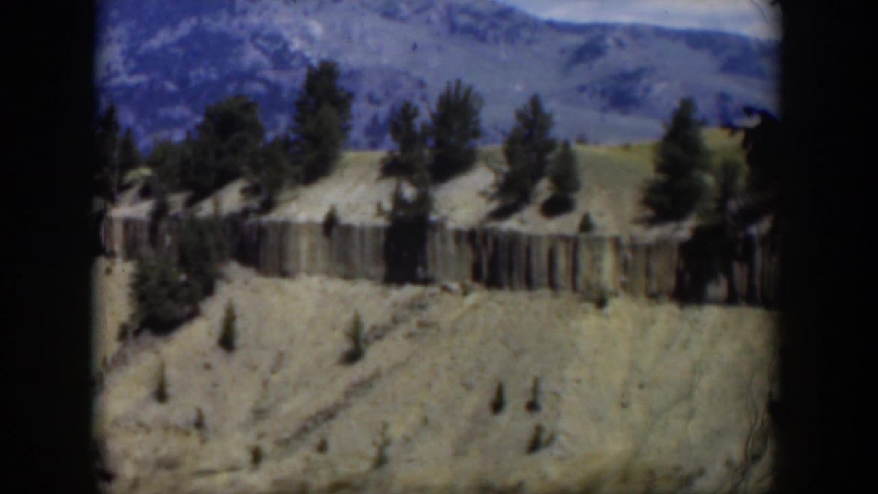 1951 Wyoming home-movie frame showing soft-focus aerial scenery with eroded hills, scattered pines, a rock band, and mountains in the background.