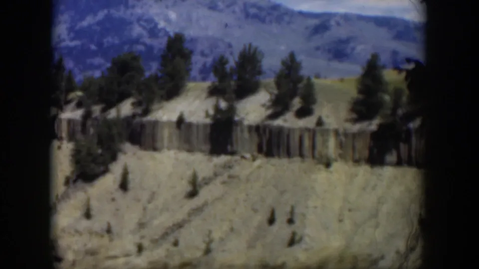 1951 Wyoming home-movie frame showing soft-focus aerial scenery with eroded hills, scattered pines, a rock band, and mountains in the background.