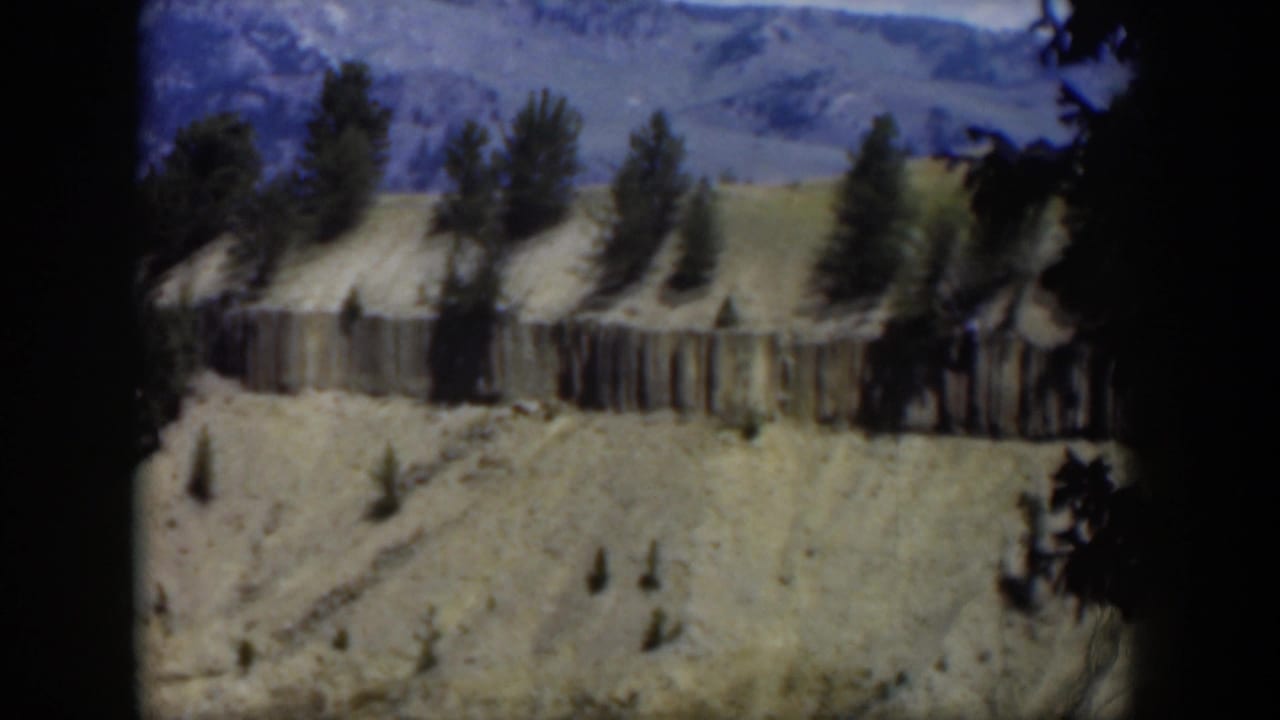 1951 Wyoming 8mm frame showing a pale ridge, vertical cliff texture, scattered pines, and blue mountain ridges from above.