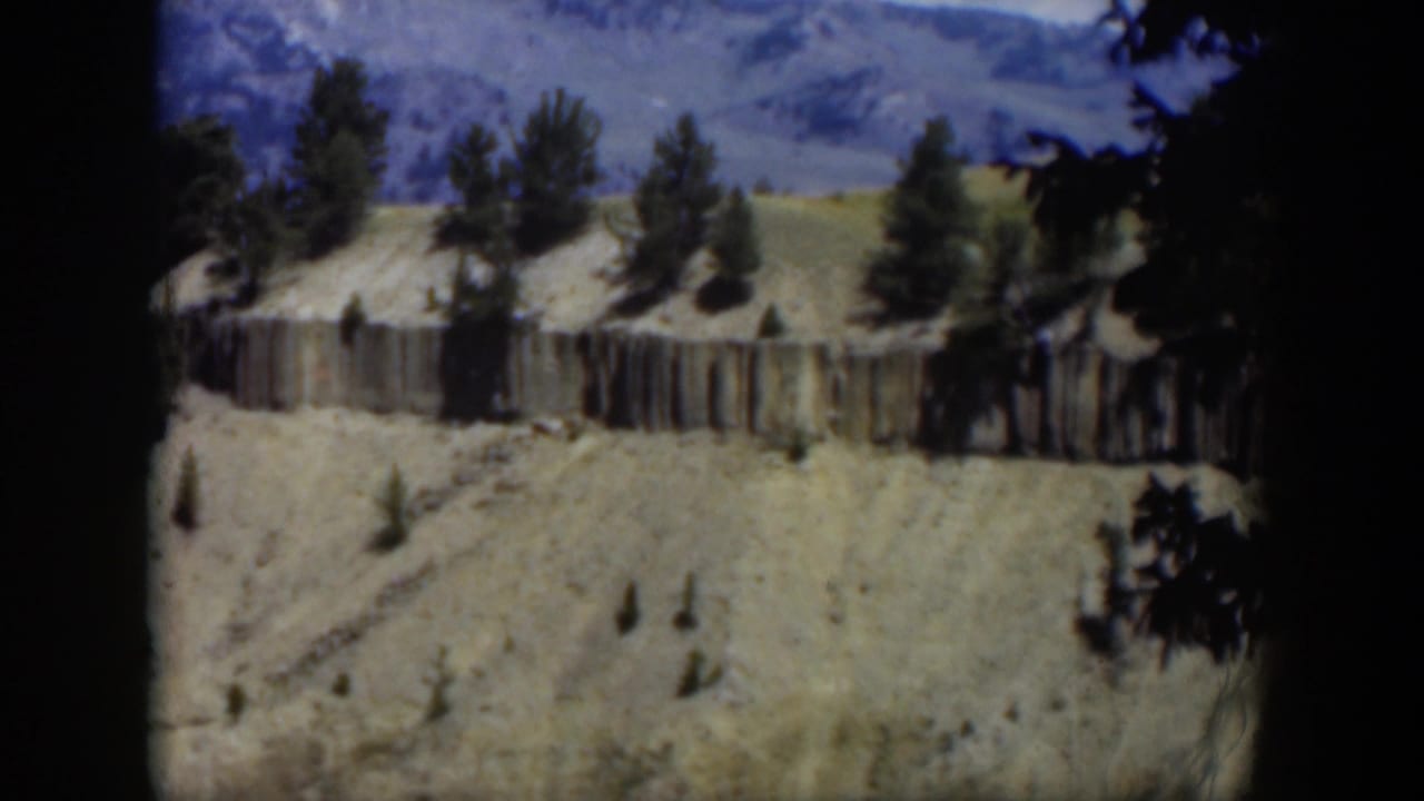 Pale mountain hillside with scattered trees, a wooden fence, and distant ridges.