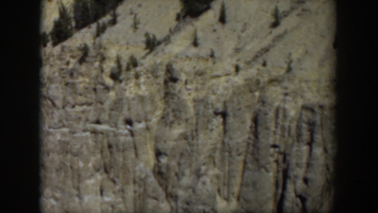 1951 Wyoming frame with a soft-focus cliff wall, sparse pines, and dry mountain terrain in faded 8mm color.