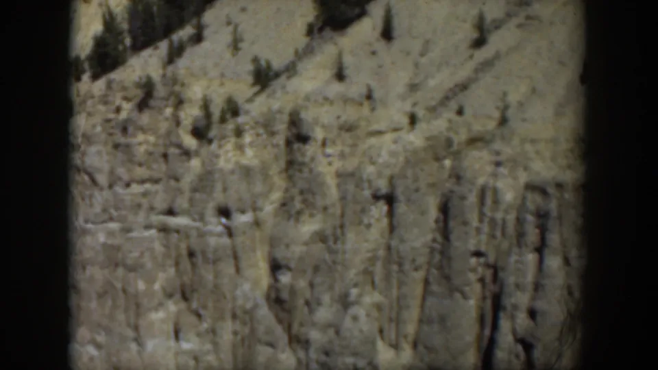1951 Wyoming frame with a soft-focus cliff wall, sparse pines, and dry mountain terrain in faded 8mm color.