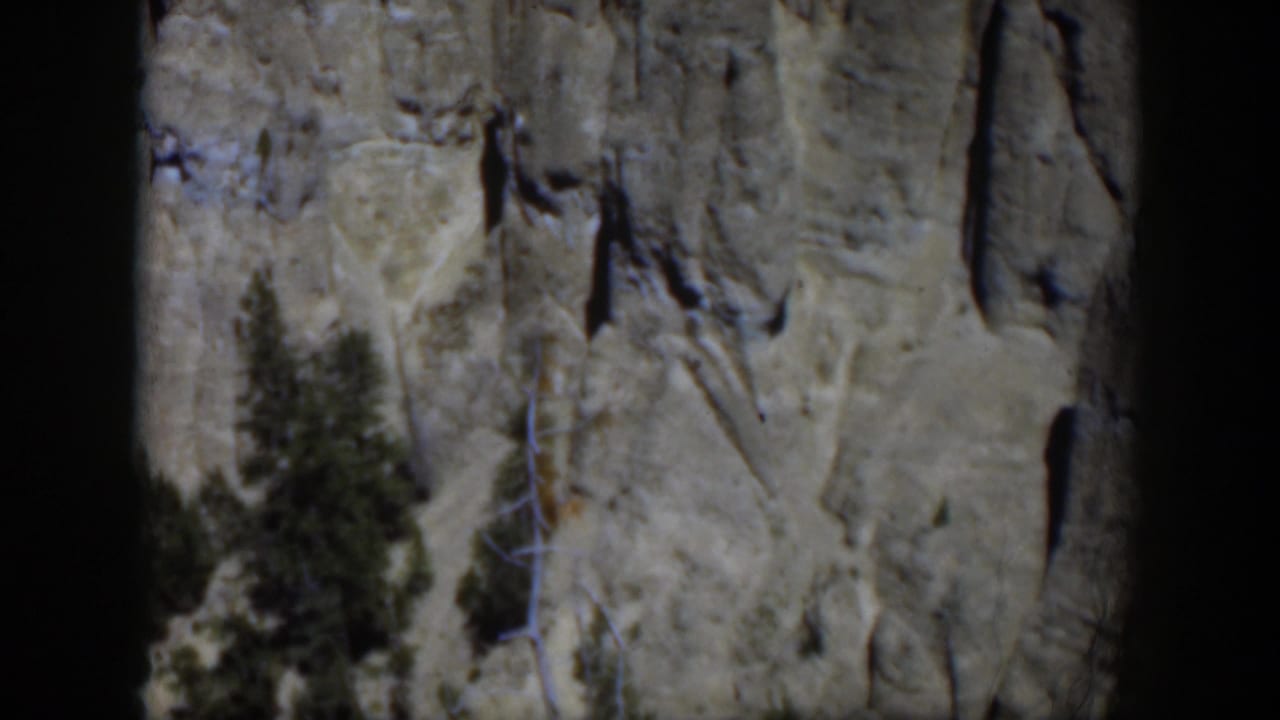 1951 Wyoming 8mm frame showing a pale gray rock wall, vertical cracks, and evergreen trees in bright daylight.