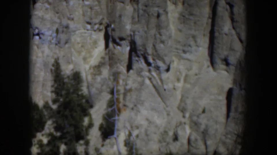 1951 Wyoming 8mm frame showing a pale gray rock wall, vertical cracks, and evergreen trees in bright daylight.