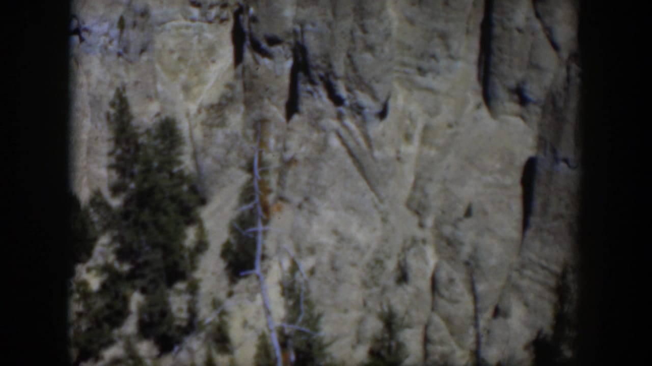 1951 Wyoming landscape frame with a gray stone wall, shadowed fissures, and sparse evergreens near the base.