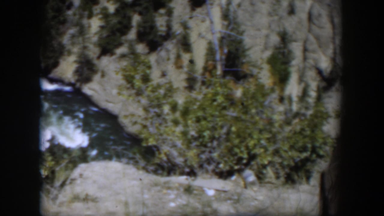 1951 Wyoming 8mm frame with a river bend below shrubs, evergreens, and pale cliff rock.