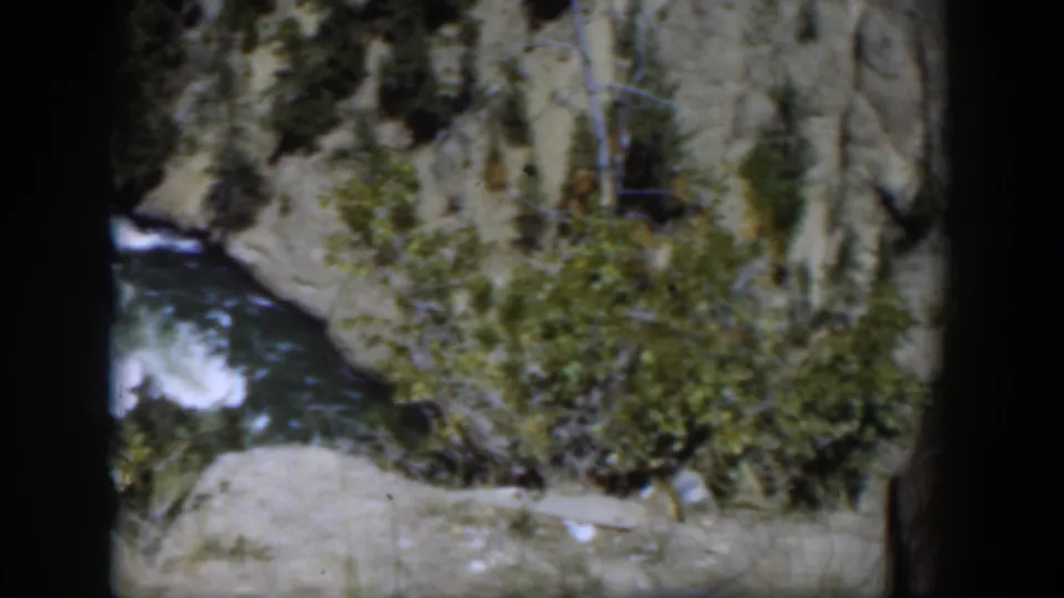 1951 Wyoming 8mm frame with a river bend below shrubs, evergreens, and pale cliff rock.