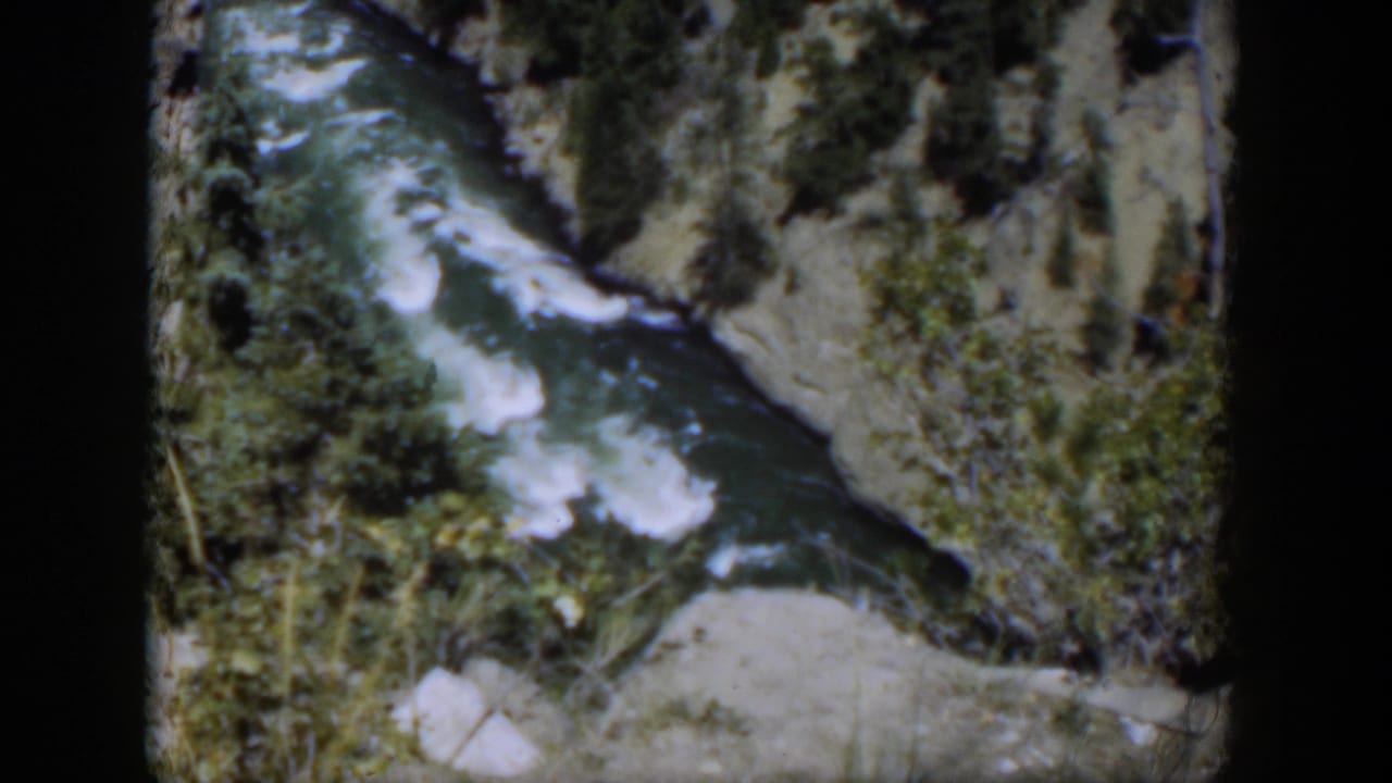 1951 Wyoming 8mm frame with a foaming river channel, pale rocks, green shrubs, and steep canyon edges.