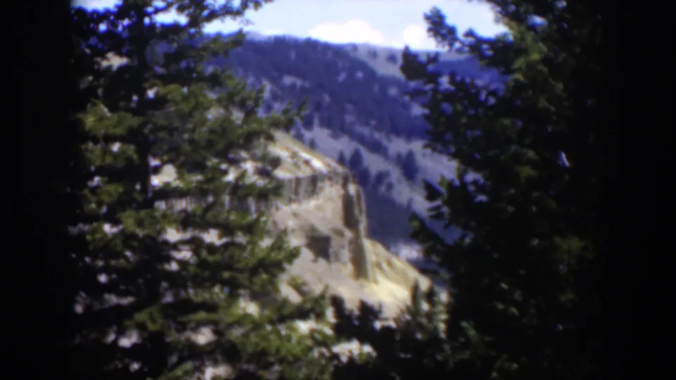 A steady 8mm view of Wyoming mountain terrain, with evergreens in the foreground and a rugged cliff beyond.