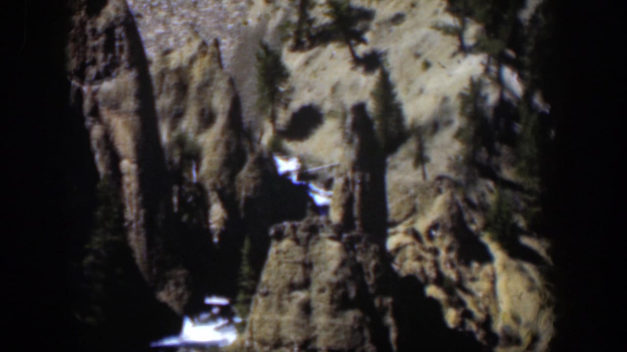 Sunlit Wyoming canyon rocks with evergreens, snow patches, and bright water below.